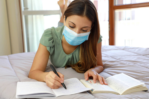 COVID-19 Πανδημία Coronavirus Πολύ κομψή μαθήτρια που φοράει χειρουργική μάσκα ξαπλωμένη στο κρεβάτι μελετώντας τα μαθήματά της κατά τη διάρκεια της καραντίνας στο σπίτι. κατ 'οίκον διδασκαλία, κοινωνική απόσταση κατά τη διάρκεια της καραντίνας, αυτο-απομόνωση, εκπαίδευση κατ' οίκον διδασκαλία έννοια. - Φωτογραφία, εικόνα