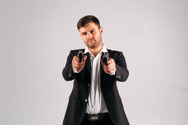 Молодой парень в черном костюме с пистолетом в руках на белом фоне в студии, он изображает охранника, телохранителя или агента
 - Фото, изображение