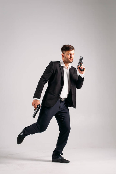 A young guy in a black suit with a gun in his hands on a white background in the studio, he depicts a security guard, bodyguard or Agent - Foto, imagen