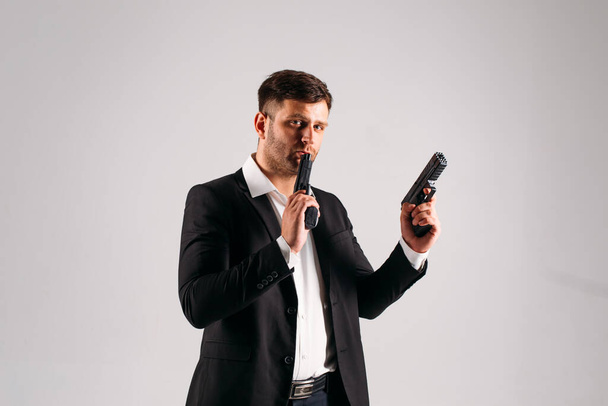 A young guy in a black suit with a gun in his hands on a white background in the studio, he depicts a security guard, bodyguard or Agent - Fotoğraf, Görsel