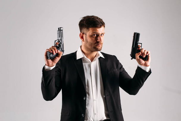 A young guy in a black suit with a gun in his hands on a white background in the studio, he depicts a security guard, bodyguard or Agent - Фото, зображення