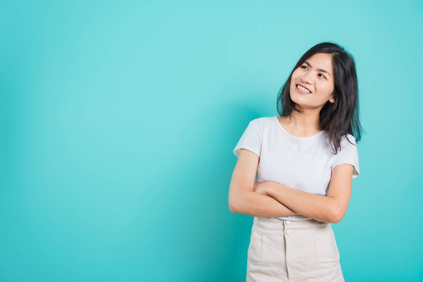 Portrait Asian beautiful happy young woman smile wear white t-shirt standing with a chest looking up away at copy space, on a blue background - Fotografie, Obrázek