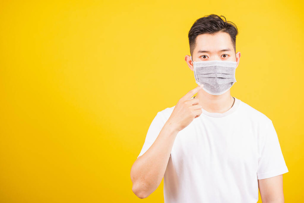 Asian young teen man wearing surgical hygienic protective face mask against coronavirus and pointing finger to mask on face, studio shot isolated white background, COVID-19 medical concept - Фото, изображение