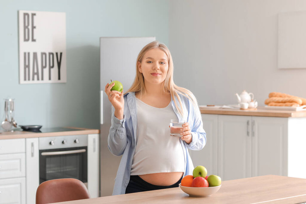 Mutfakta elma ve bardak su taşıyan hamile bir kadın. - Fotoğraf, Görsel