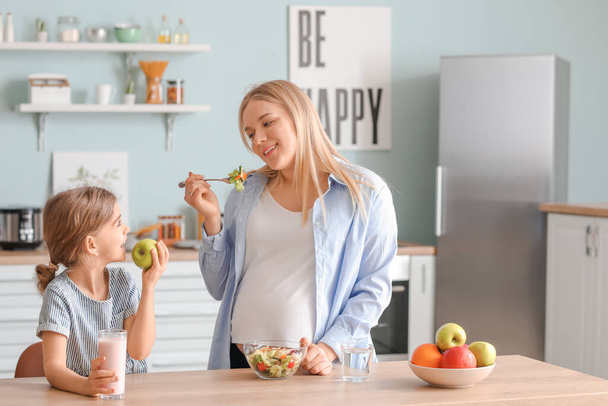 Беременная женщина со своей маленькой дочерью, которая ест на кухне
 - Фото, изображение