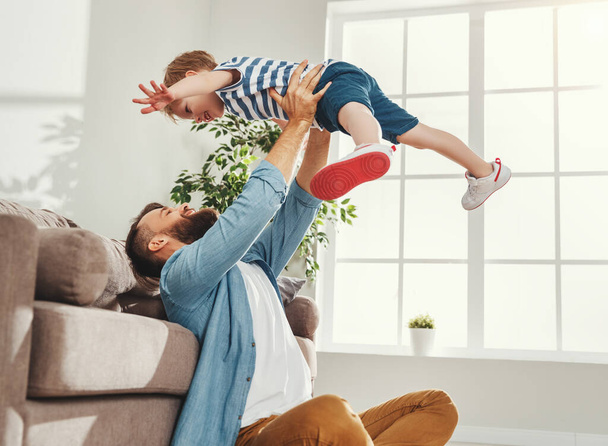Радостный молодой человек в повседневной одежде сидит на полу и бросает счастливого маленького сына, веселясь вместе в уютной гостиной в доме
 - Фото, изображение