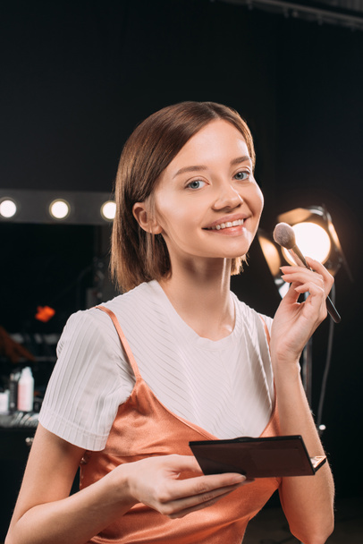 Beautiful woman smiling at camera while holding cosmetic brush and set of blush in photo studio  - Valokuva, kuva