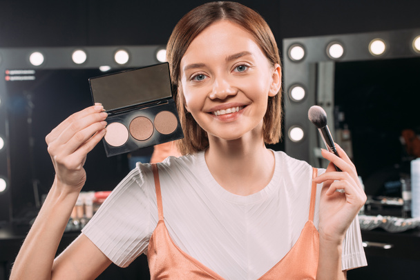 Hermosa mujer sonriente sosteniendo cepillo cosmético y maquillaje conjunto en el estudio de fotos
  - Foto, imagen