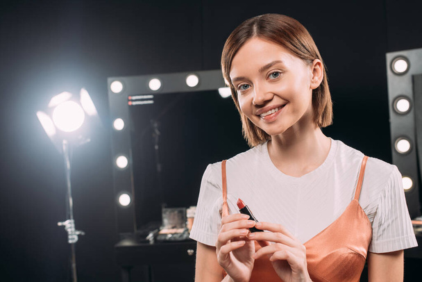 Beautiful smiling woman holding red lipstick in photo studio  - Zdjęcie, obraz