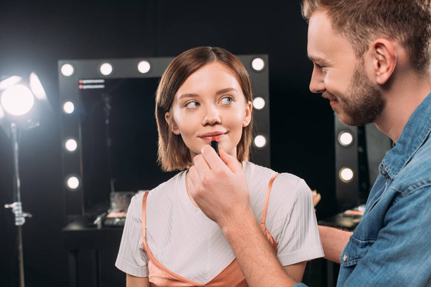 Smiling makeup artist applying red lipstick on attractive young model in photo studio  - Photo, Image
