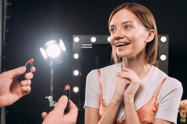 Makeup artist holding lipsticks near positive model in photo studio  - Φωτογραφία, εικόνα