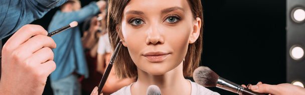 Panoramic orientation of makeup artists holding cosmetic brushes near beautiful model in photo studio  - 写真・画像