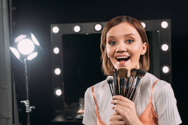 Cheerful model with cosmetic brushes looking at camera in photo studio  - Фото, зображення