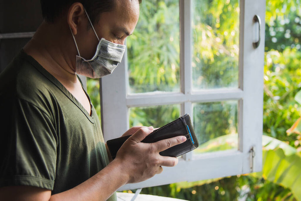 Közelkép ázsiai férfi zöld alkalmi ing visel influenza maszk nézi a pénztárcáját, ő áll mellett egy ablak zöld természet elmosódott háttér. - Fotó, kép