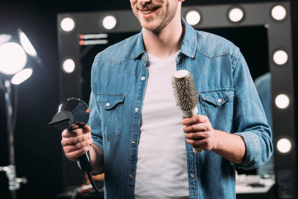 Обрізаний вид усміхненого візажиста, який тримає щітку для волосся та фен у фотостудії
  - Фото, зображення