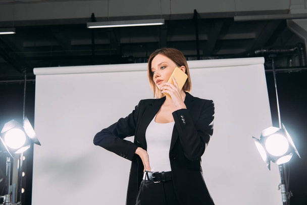 Low angle view of beautiful woman in formal wear talking on smartphone in photo studio  - Fotografie, Obrázek