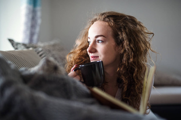Young woman relaxing indoors at home with book and cup of coffee. - Valokuva, kuva