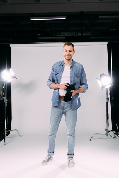 Handsome photographer holding digital camera and smiling at camera in photo studio  - Фото, изображение