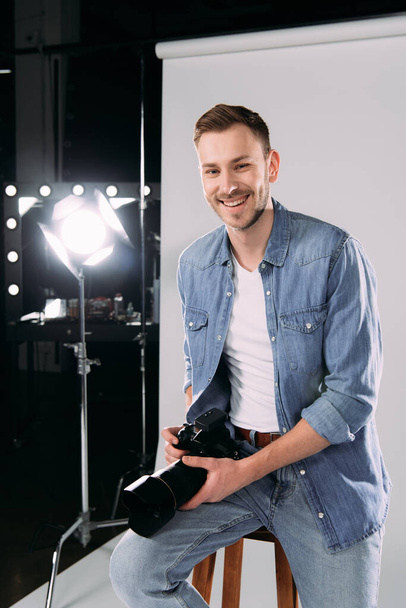 Χαμογελώντας φωτογράφος κρατώντας ψηφιακή φωτογραφική μηχανή κοντά floodlight στο στούντιο φωτογραφιών  - Φωτογραφία, εικόνα