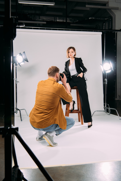 Selective focus of photographer working with stylish model on chair in photo studio  - 写真・画像