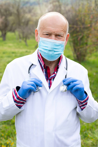 Portrait of senior male doctor with uniform and surgical mask posing outdoors - Foto, afbeelding