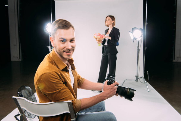 Lächelnder Fotograf blickt in die Kamera, während das Model einen Blumenstrauß im Fotostudio hält  - Foto, Bild
