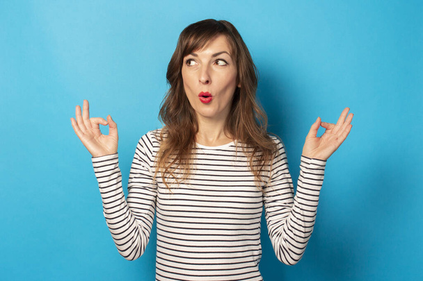 Portret van een jonge vriendelijke vrouw met een verrast gezicht in een casual t-shirt maakt een zen gebaar op een geïsoleerde blauwe achtergrond. Emotioneel gezicht. Ontspanning, meditatie, stress. - Foto, afbeelding