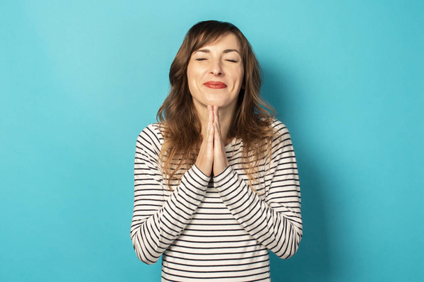 Young friendly woman with a smile in a casual t-shirt making prayer gesture with closed eyes on an isolated background. Emotional face. To ask for something from the universe or from God, make a wish. - Фото, изображение