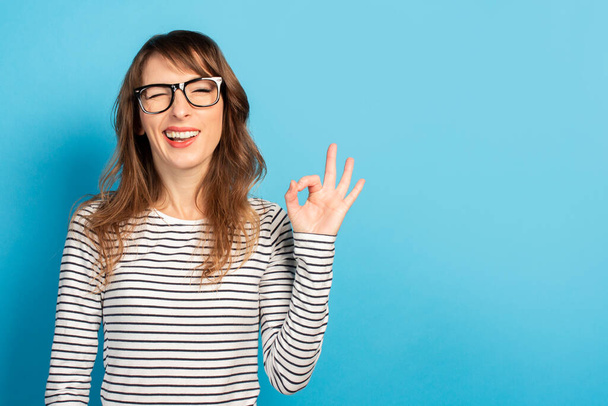 Retrato de uma jovem mulher amigável com um sorriso em uma camiseta casual e óculos faz um gesto ok em um fundo de luz isolado. Cara emocional. O gesto está bem, está tudo bem.
. - Foto, Imagem