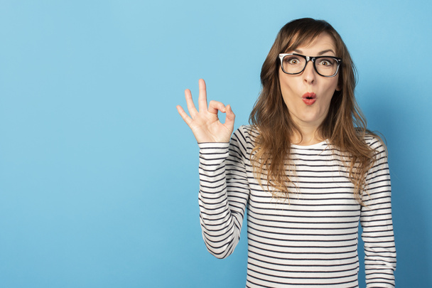 Portrait of a young friendly woman with a surprised face in a casual t-shirt and glasses makes an okay gesture on an isolated light background. Emotional face. Gesture is all right, it's OK. - Foto, Imagem