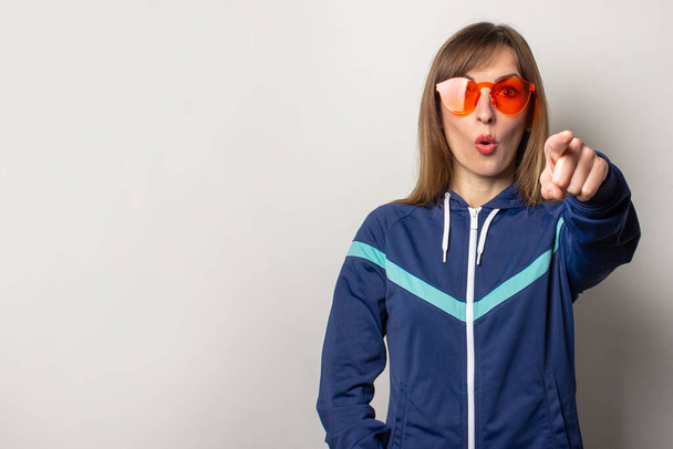 giovane donna sorpresa in una giacca sportiva anni '90 e occhiali rossi su uno sfondo leggero. Il modello punta un dito dritto davanti a sé e guarda la fotocamera. Concetto di gesti, stile, moda, sport
.  - Foto, immagini