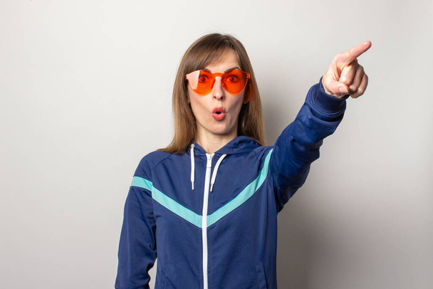 έκπληκτη νεαρή γυναίκα με αθλητικό μπουφάν της δεκαετίας του '90 και κόκκινα γυαλιά σε ελαφρύ φόντο. Το μοντέλο δείχνει ένα δάχτυλο ευθεία και κοιτάζει την κάμερα. Έννοια χειρονομίες, στυλ, μόδα, αθλητισμός. - Φωτογραφία, εικόνα