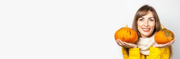 boldog fiatal lány sárga kabátban két sütőtököt tart könnyű háttérben. Halloween koncepció, ősz, ünneplés. Zászlós. - Fotó, kép