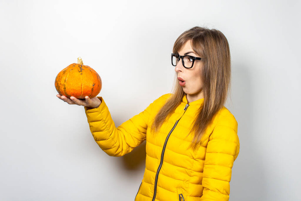 a menina jovem em uma jaqueta amarela mantém uma abóbora com uma cara assustada em um contexto leve. Conceito de Halloween, outono, celebração. - Foto, Imagem