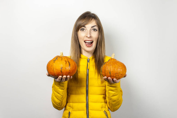счастливая молодая девушка в желтой куртке держит две тыквы на светлом фоне. Хэллоуин концепции, осень, празднование.  - Фото, изображение