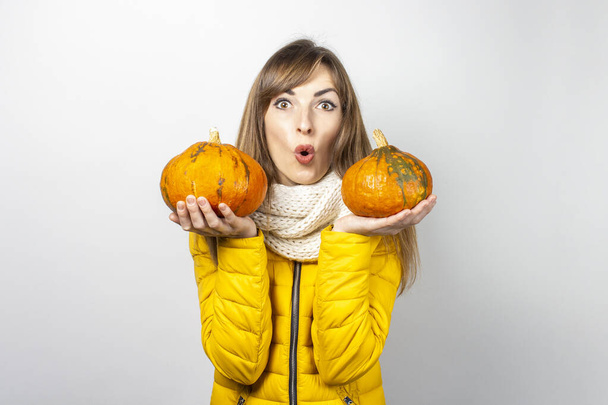 шокированная молодая девушка в жёлтой куртке держит две тыквы на светлом фоне. Хэллоуин концепции, осень, празднование.  - Фото, изображение