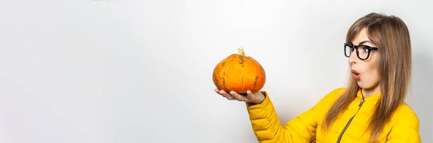 молодая девушка в жёлтой куртке держит тыкву с испуганным лицом на светлом фоне. Концепция Хэллоуина, осень, праздник. Баннер
. - Фото, изображение