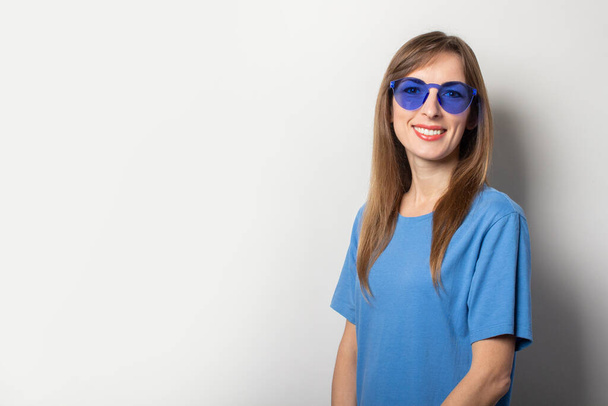 Portret van een jonge vriendelijke vrouw in casual blauw t-shirt en blauwe bril glimlachend op een geïsoleerde lichte achtergrond. Emotioneel gelaat. - Foto, afbeelding
