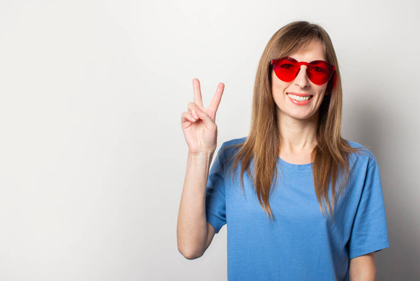 Portrét mladé přátelské ženy s úsměvem v neformálním modrém tričku, s červenými brýlemi, udělá pozdrav, dva prsty na osamělém světlém pozadí. Emocionální tvář. - Fotografie, Obrázek