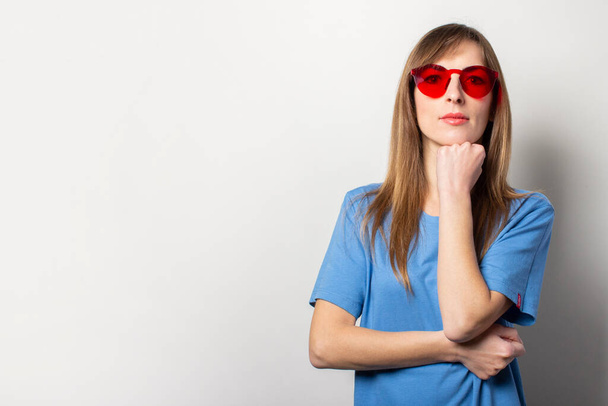 Portrait of a young friendly woman in a casual blue t-shirt, red glasses, holds her hand on her chin on an isolated light background. Emotional face. Gesture to think, plan. - Photo, Image
