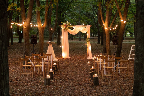 Bruidsboog in het bos met gloeilampen. Mooie rustieke bruiloft in het bos. Zitplaatsen voor gasten op een prachtige bruiloft met gloeilampen in het bos. - Foto, afbeelding