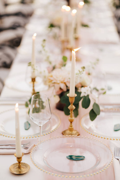 Wedding dinner table at reception. Beautiful white delicate candles burn in metal candlesticks, against background of white and pink tablecloth, flower arrangements and glass plates with gold beads - Photo, Image
