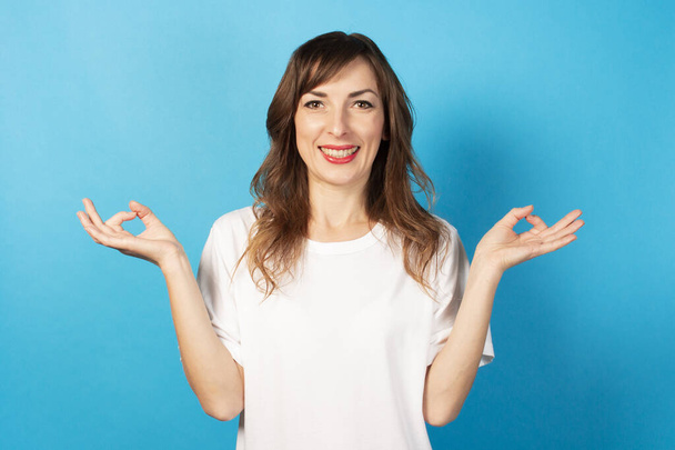 Портрет молодої дружньої жінки в повсякденній футболці з жестом медитації та посмішкою на ізольованому синьому фоні. Емоційне обличчя. Жест для розслаблення
. - Фото, зображення