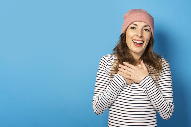 Cute young woman with a surprised face in a hat and striped sweater laid two palms on her chest against a blue background. Emotional face. Gesture of excitement, joy, surprise. - Photo, Image