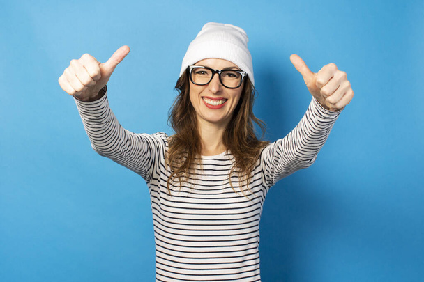 Portrait of a Cute Young woman in glasses, a hat and a jacket holds her hands in front of herself with a gesture OK, everything is fine, thumb up on a blue background. Emotional face. - Photo, Image