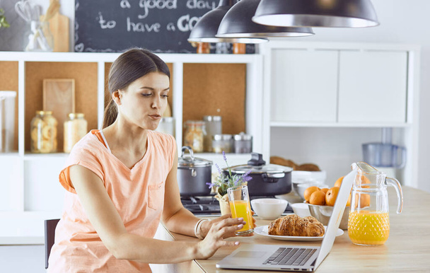 Uśmiechający się ładna kobieta, patrząc na telefon komórkowy i trzymając szklankę soku pomarańczowego, jedząc śniadanie w kuchni - Zdjęcie, obraz