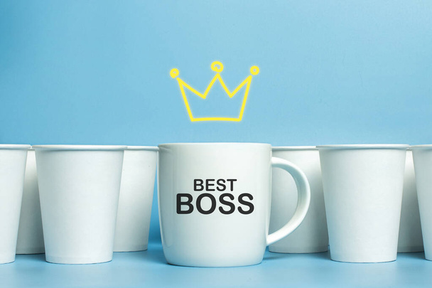 白いカップの上に王冠が描かれ、青い背景に白い紙コップで囲まれています。コンセプトボス、ユニークでフレンドリーなチーム。コピースペース。追加テキスト｜Best Boss. - 写真・画像