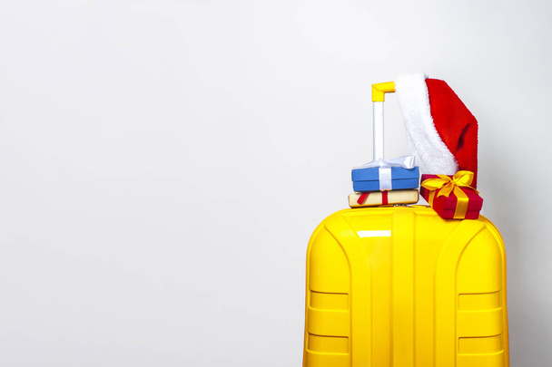 On a yellow plastic suitcase are gifts and wearing a red Santa Claus hat on a light background. - Photo, Image