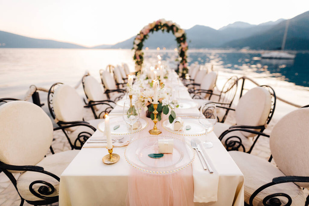 Крупный план свадебного ужина. Стол стоит на пляже с видом на горы на закате. Металлические кованые стулья, горящие свечи, свадебная арка из цветов, скатерть кремового цвета
. - Фото, изображение