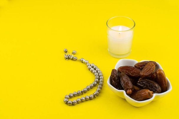 Festiwal Ramadan Kareem. Zbliżenie suszonych dat owoców w misce ze świecą i różańcem na żółtym tle. Przestrzeń kopiowania - Zdjęcie, obraz